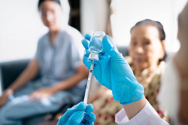 Image focused on a healthcare worker's hands prepping a vaccine for their patient, elderly Asian woman, waiting in the background