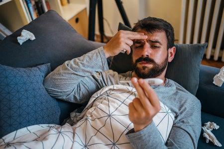 Sick man, wrapped in a blanket, is measuring a temperature while lying in bed.