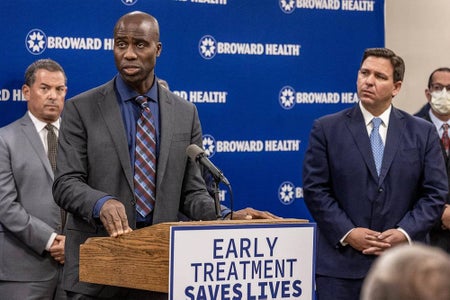 Florida Surgeon General Joseph Ladapo, left, speaks at a podium with a sign on front that reads "EARLY TREATMENT SAVES LIVES," during a press conference at Broward Health Medical Center on Jan. 3, 2022, as Gov. Ron DeSantis, right, listens