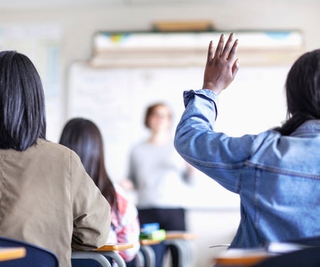 A student in a classroom, seen from behind, raises a hand.