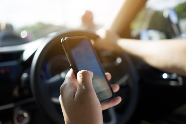 A person driving an automobile, one hand on the steering wheel, one hand holding up a smartphone