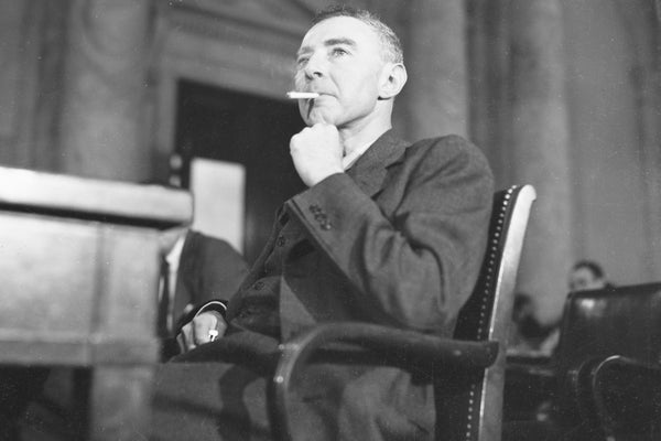 Archival image of J. Robert Oppenheimer smoking a cigarette while sitting behind desk, testifying before congress.