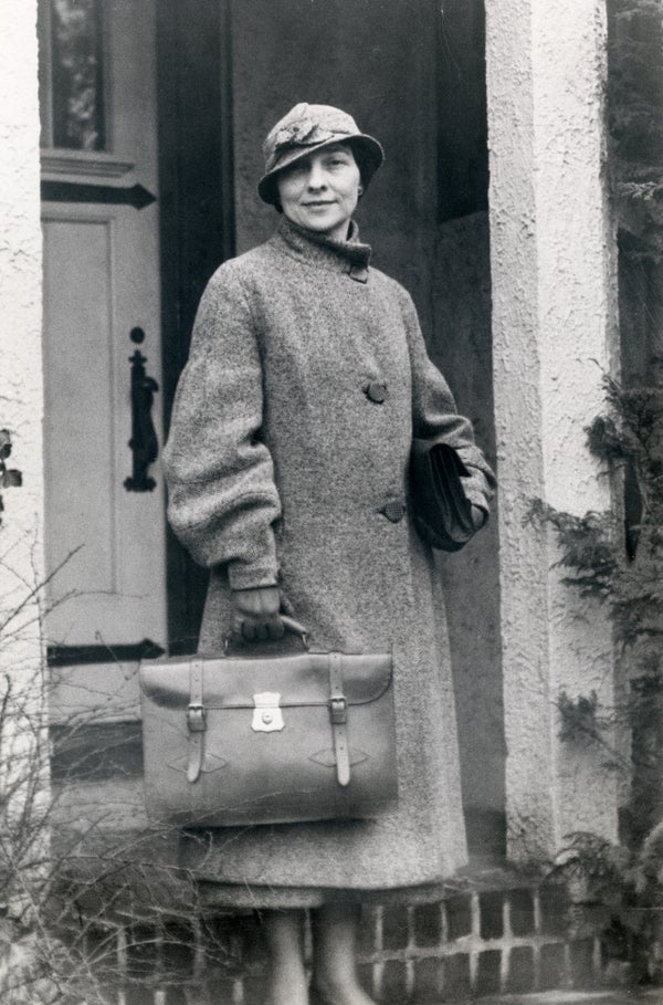 A woman in a trench coat holding a briefcase stands looking toward the camera