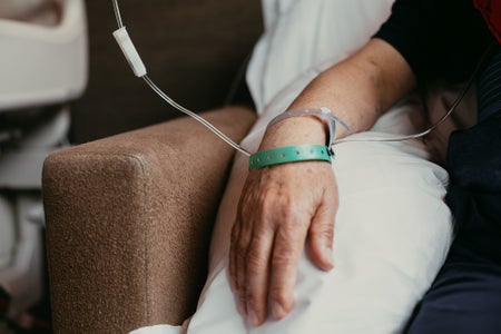 Close-up of an elderly patient's arm and hand, wearing medical bracelets, receiving treatment in a hospital while sitting in an upholstered chair