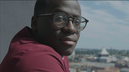 A man with glasses stands outside and looks into the camera over his right shoulder