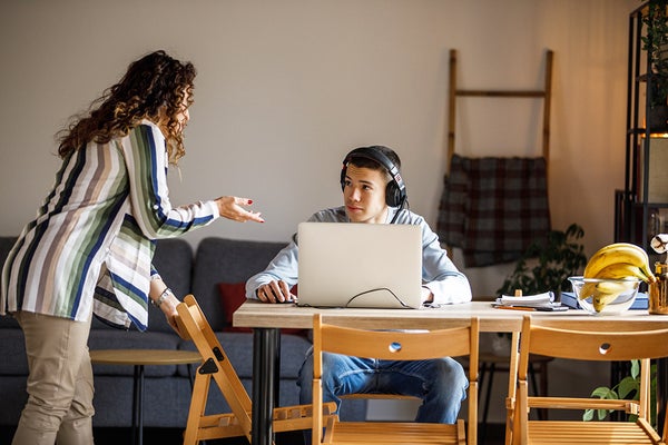 Mother interacting with teenage boy who is woking on laptop with headphones