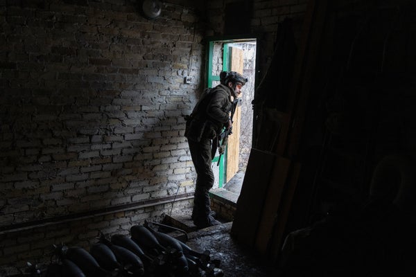 An armed soldier peers out from the darkened doorway of a brick structure