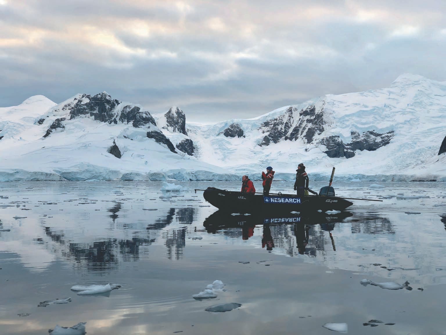 Three people in a small whale tagging boat shown in Antarctic water with snow covered land in background.
