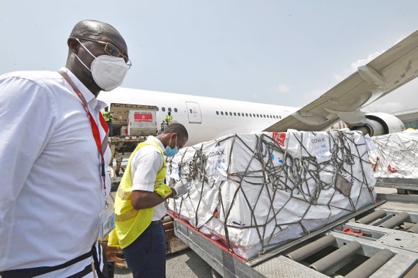 Men with masks stand near a pallet wrapped in plastic that just came off an airplane, which stands in the background.