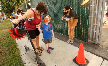 Little boy with face mask looks up at woman who is holding a scooter as aide looks on.