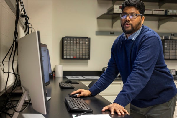 Physicist Ranga Dias in blue sweater standing at desk in front of a computer screen in his lab.