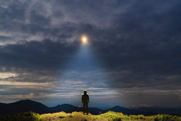 A man standing in a field on a cloudy night looking up in wonder at a very bright light peeking through the clouds.