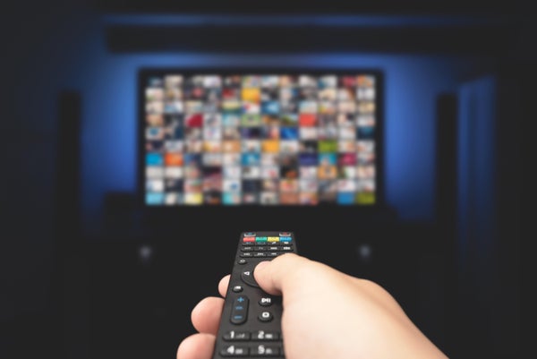 Person with remote control in hand pointing at TV screen in a dark room.