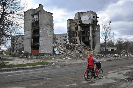 Woman in red jacket with red bike walks past buildings in rubble