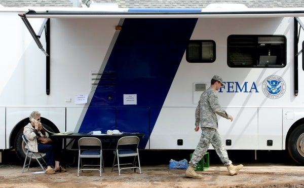 Woman using a phone in front of a FEMA truck; a soldier also walks in front of the truck.