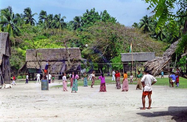A group of people in tropical garb surrounded by mostly thatch-roofed one-story buildings.