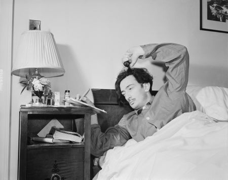 A man lays in bed writing on a note pad and twisting his hair with his fingers