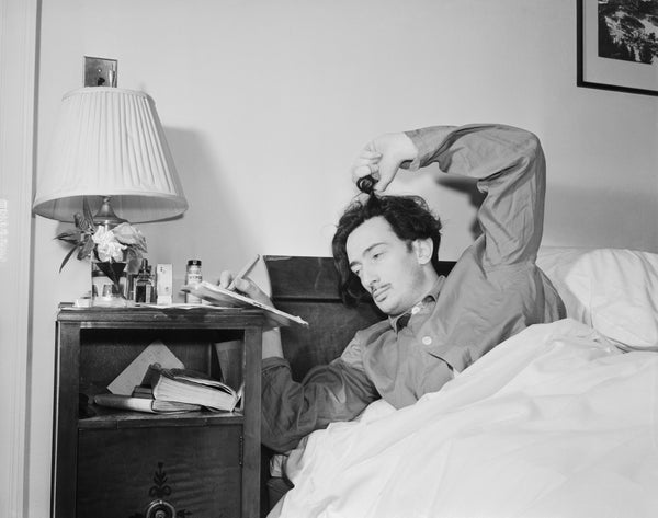A man lays in bed writing on a note pad and twisting his hair with his fingers