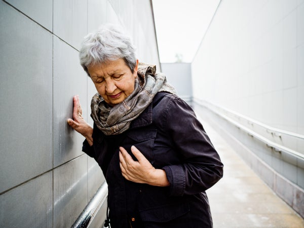 An older woman leans against a wall in obvious discomfort.