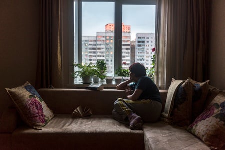 A seated woman gazes from her darkened living room onto a cityscape.