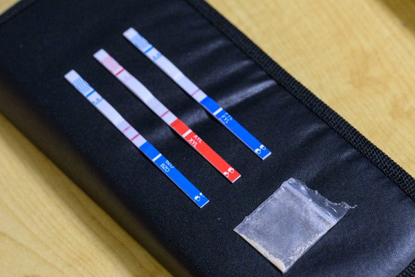 Three test strips shown against a black background, next to a small packet of heroin.