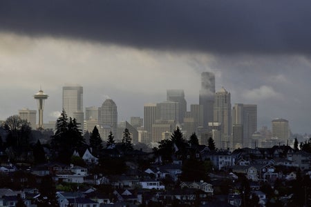 Seattle skyline seen between overcast clouds above and, in the foreground, a hilly residential neighborhood in shadow