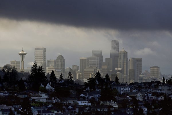 Seattle skyline seen between overcast clouds above and, in the foreground, a hilly residential neighborhood in shadow
