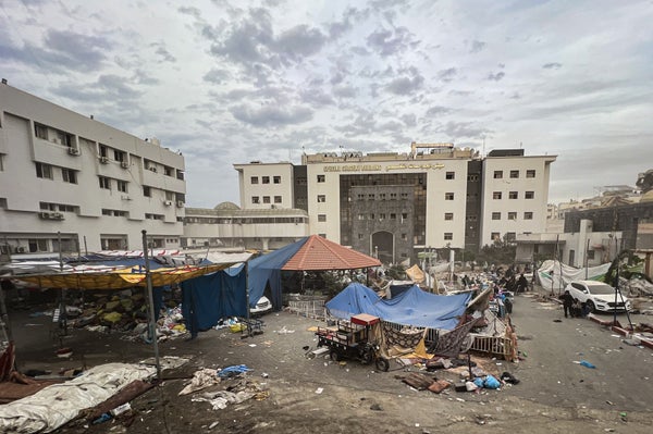 Photograph of the exterior of bombed-out Al-Shifa Hospital in Gaza City surrounded by scattered debris, tarps and tents