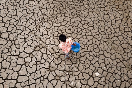 Aerial of a man carrying a blue bucket walking on cracked dry soil to fetch water.