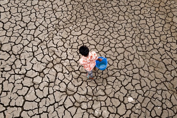 Aerial of a man carrying a blue bucket walking on cracked dry soil to fetch water.