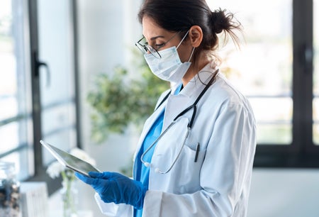 A female physician in scrubs and a mask checks her tablet.