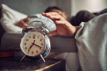 A young man reaching for his alarm clock after waking up in bed at home.