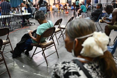 2 elderly women - one with a white hair bow - sitting in chairs with others in the background of a cooling facility